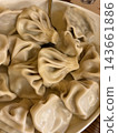 Uncooked folded dumplings with twisted pleats and smooth glossy dough fill a shallow bowl, viewed closely beside a wooden surface 143661886