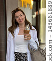 Smiling woman holds a chocolate topped ice cream cone beside a building, wearing sunglasses, white clothing, jewelry, and carrying a handbag 143661889