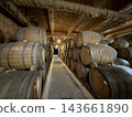 Wooden barrels are stacked along both sides of a narrow concrete walkway beneath exposed pipes and a textured low ceiling 143661890