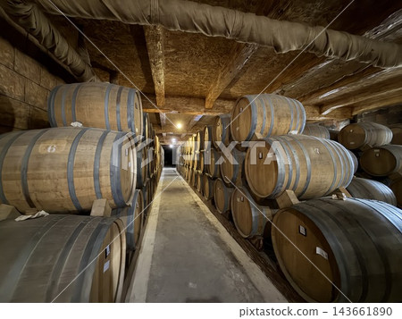 Wooden barrels are stacked along both sides of a narrow concrete walkway beneath exposed pipes and a textured low ceiling 143661890