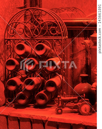 Red lighting reveals an ornate metal rack filled with rolled objects, a bottle, and a small toy tractor on a wooden shelf Red lighting reveals an ornate metal rack filled with rolled objects, a bottle, and a small toy tractor on a wooden shelf 143661891