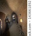 Dim underground passage with rough earthen walls, stacked stones, arched openings, and warm light visible through doorways in a historic subterranean interior 143661892