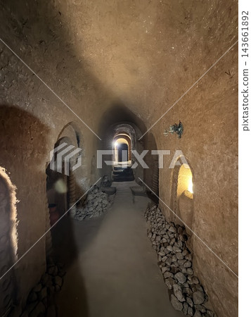 Dim underground passage with rough earthen walls, stacked stones, arched openings, and warm light visible through doorways in a historic subterranean interior 143661892