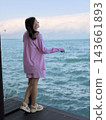 Woman wearing pink striped clothing stands on a dark wooden deck beside blue ocean water, with distant clouds and gentle waves beyond 143661893