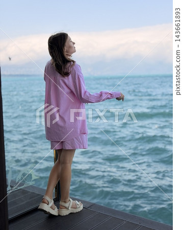 Woman wearing pink striped clothing stands on a dark wooden deck beside blue ocean water, with distant clouds and gentle waves beyond Woman wearing pink striped clothing stands on a dark wooden deck beside blue ocean water, with distant clouds and gentle waves beyond 143661893