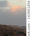 Soft pink clouds glow above misty rugged hills, with sparse vegetation and layered distant ridges forming a muted arid mountain landscape 143661894
