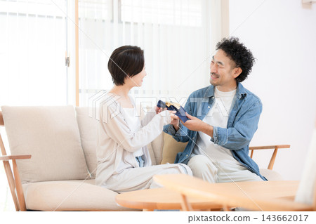 一位日本妻子將禮物送給坐在客廳沙發上的丈夫;丈夫接過禮物時,這對恩愛的夫妻臉上露出了笑容。 一位日本妻子將禮物送給坐在客廳沙發上的丈夫;丈夫接過禮物時,這對恩愛的夫妻臉上露出了笑容。 143662017