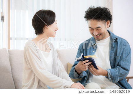 A Japanese wife presents a gift to her husband as he sits on the sofa in their living room; the loving couple smiles as the husband accepts the gift. A Japanese wife presents a gift to her husband as he sits on the sofa in their living room; the loving couple smiles as the husband accepts the gift. 143662019