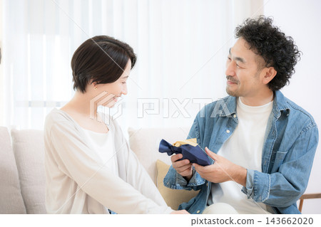 A Japanese wife presents a gift to her husband as he sits on the sofa in their living room; the loving couple smiles as the husband accepts the gift. 143662020