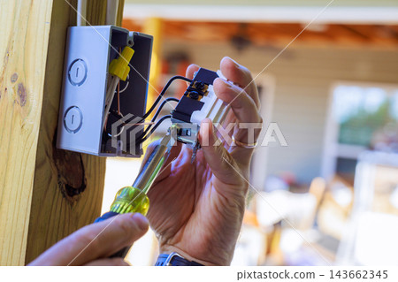 An electrician uses screwdriver to test wiring in an open electrical box inside building. 143662345