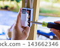 Contractor hands fit an outlet box to wooden frame while screwdriver tightens screw outdoors 143662346