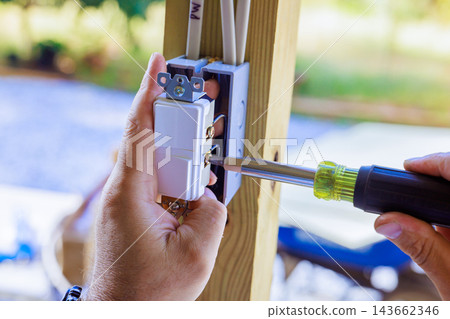 Contractor hands fit an outlet box to wooden frame while screwdriver tightens screw outdoors Contractor hands fit an outlet box to wooden frame while screwdriver tightens screw outdoors 143662346