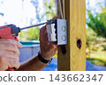 Worker uses power screwdriver to attach an electrical switch box to wooden post outdoors 143662347