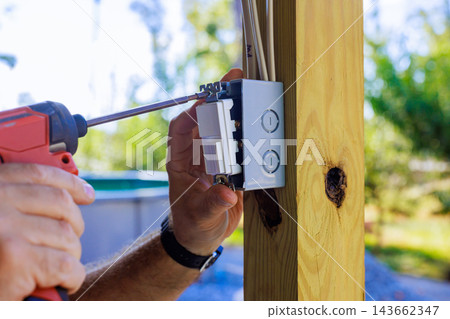 Worker uses power screwdriver to attach an electrical switch box to wooden post outdoors Worker uses power screwdriver to attach an electrical switch box to wooden post outdoors 143662347