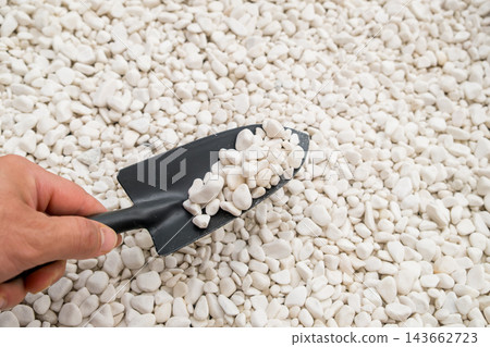 Scooping small white pebbles using a black hand trowel from a large pile for gardening or landscaping purposes. Scooping small white pebbles using a black hand trowel from a large pile for gardening or landscaping purposes. 143662723