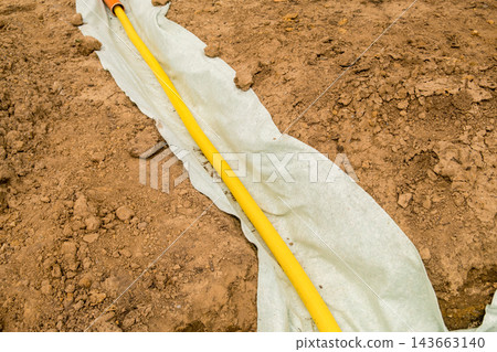 In a prepared landscaping trench, a yellow drainage hose is positioned on protective fabric, surrounded by soil and organized materials. In a prepared landscaping trench, a yellow drainage hose is positioned on protective fabric, surrounded by soil and organized materials. 143663140