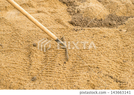 A rake is seen leveling fine sand at a construction site, ensuring an even surface. The focus is on the meticulous action of smoothing the sand for landscaping purposes. 143663141