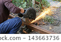 A man in work clothes is cutting a rusty metal profile pipe outdoors using a chop saw. Bright sparks of molten metal fly in all directions from beneath the rotating abrasive disc. 143663736