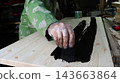 A craftsman wearing a protective glove paints a smooth wooden board with black paint. The process of staining the wood using a foam sponge takes place on a workbench in a workshop. 143663864