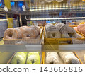 A variety of colorful donuts dusted with powdered sugar are neatly arranged in rows within woven baskets inside a glass bakery display case at a cafe or pastry shop. 143665816