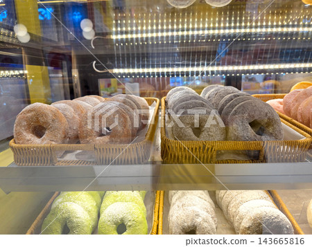 A variety of colorful donuts dusted with powdered sugar are neatly arranged in rows within woven baskets inside a glass bakery display case at a cafe or pastry shop. A variety of colorful donuts dusted with powdered sugar are neatly arranged in rows within woven baskets inside a glass bakery display case at a cafe or pastry shop. 143665816
