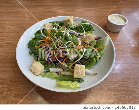 A fresh garden salad with mixed greens, croutons, and sunflower sprouts in a white bowl, served with creamy dressing on a wooden table. 143665818