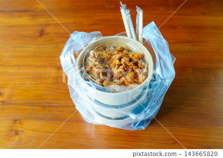 A Korean takeaway food order of spicy meat and rice in paper bowls, packed in a clear plastic bag with chopsticks on a wooden table. Represents food delivery or a convenient office lunch. A Korean takeaway food order of spicy meat and rice in paper bowls, packed in a clear plastic bag with chopsticks on a wooden table. Represents food delivery or a convenient office lunch. 143665820