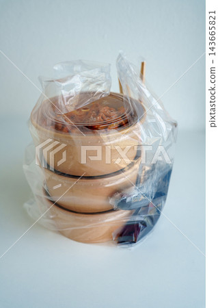Three brown kraft paper takeout food bowls with clear lids inside a plastic bag on a white background. Includes chopsticks and condiments for a meal delivery or restaurant packaging concept. 143665821