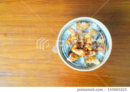 Top view of an Asian-style takeout meal with fried chicken over white rice in a round disposable container. The bowl is topped with sesame seeds and sits on a wooden table with copy space. Top view of an Asian-style takeout meal with fried chicken over white rice in a round disposable container. The bowl is topped with sesame seeds and sits on a wooden table with copy space. 143665825