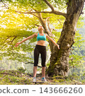 Athletic young woman standing with arms open wide and looking up towards the sky under a majestic green tree in a scenic mountain forest environment 143666260