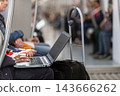Close-up of a commuter typing on a laptop computer balanced on his lap while sitting in a public train car, with a rolling luggage bag resting right beside him 143666262