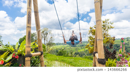 Happy sporty male traveller swinging on wooden swing, enjoying summer vacation in pristine green nature at Jatiluwih rice terraces on Bali, Indonesia. Freedom and life enjoying concept. Happy sporty male traveller swinging on wooden swing, enjoying summer vacation in pristine green nature at Jatiluwih rice terraces on Bali, Indonesia. Freedom and life enjoying concept. 143666266