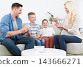 A cheerful lifestyle photo of a family of four sitting on a couch in their living room, playing a card game together. The mother, father, and two young sons are holding playing cards 143666271