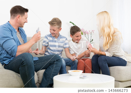 A cheerful lifestyle photo of a family of four sitting on a couch in their living room, playing a card game together. The mother, father, and two young sons are holding playing cards 143666273