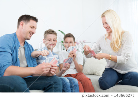 A cheerful lifestyle photo of a family of four sitting on a couch in their living room, playing a card game together. The mother, father, and two young sons are holding playing cards 143666275