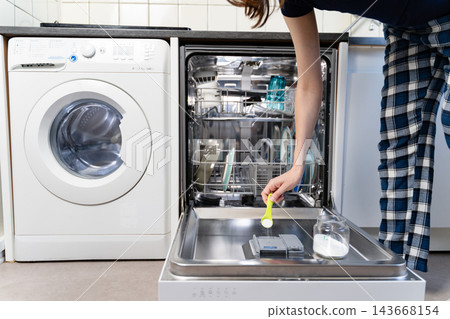 Woman adding homemade dishwasher detergent with a measuring spoon to an open dishwasher beside a washing machine in a bright modern laundry room. Woman adding homemade dishwasher detergent with a measuring spoon to an open dishwasher beside a washing machine in a bright modern laundry room. 143668154