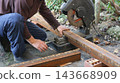 A worker tightens the screw clamp on a chop saw, securely holding a rusty profile pipe in place before cutting. The process of preparing the metal workpiece for cutting takes place outdoors 143668909