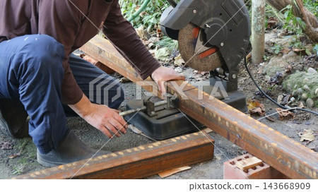 A worker tightens the screw clamp on a chop saw, securely holding a rusty profile pipe in place before cutting. The process of preparing the metal workpiece for cutting takes place outdoors A worker tightens the screw clamp on a chop saw, securely holding a rusty profile pipe in place before cutting. The process of preparing the metal workpiece for cutting takes place outdoors 143668909