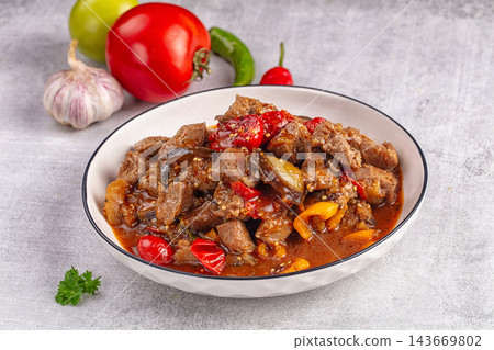 Hearty beef stew with eggplant, red peppers, and tomato sauce in white bowl, fresh garlic and parsley garnish on gray stone background 143669802