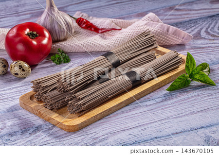 Wooden cutting board holding bundles of uncooked soba noodles beside fresh tomato, garlic, basil, quail eggs, and red chili pepper on rustic painted wood surface 143670105
