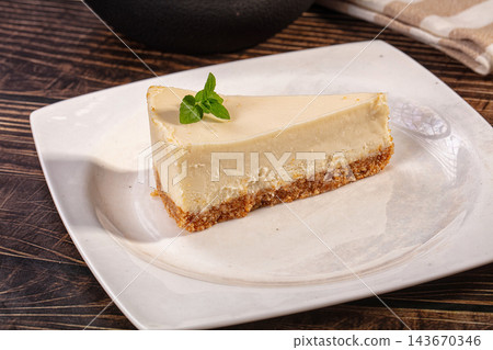 Slice of classic cheesecake with biscuit crust and fresh mint leaf served on white plate over rustic dark wooden table, dessert photography Slice of classic cheesecake with biscuit crust and fresh mint leaf served on white plate over rustic dark wooden table, dessert photography 143670346
