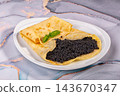 White ceramic plate on grey marble with folded golden crepes filled with glossy black caviar, basil garnish, gourmet delicacy close up 143670347