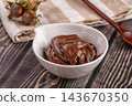 Wooden spoon and fresh hazelnuts beside white ceramic bowl of creamy chocolate spread on rustic wooden table, sweet dessert topping 143670350