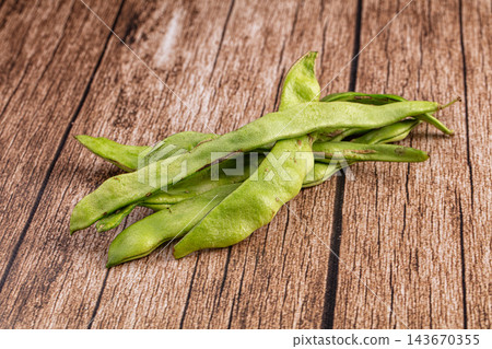 Raw green bean pods scattered on rustic brown wooden table, fresh flat runner beans with natural texture, healthy vegetable food ingredient Raw green bean pods scattered on rustic brown wooden table, fresh flat runner beans with natural texture, healthy vegetable food ingredient 143670355
