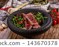 Sliced medium rare beef steak with arugula and cherry tomatoes in black stone bowl on rustic wooden table with garlic, chili and linen cloth, gourmet meat dish 143670378
