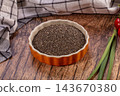 Raw chia seeds filling orange ceramic bowl on rustic wood table with checkered linen, cherry tomatoes and green onion, healthy superfood seeds 143670380