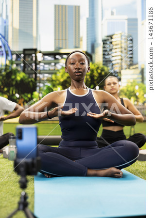 Yoga instructor woman demonstrates controlled breathing movement for smartphone vlog recording, session with seated class participants on yoga mats in urban park. Yoga instructor woman demonstrates controlled breathing movement for smartphone vlog recording, session with seated class participants on yoga mats in urban park. 143671186