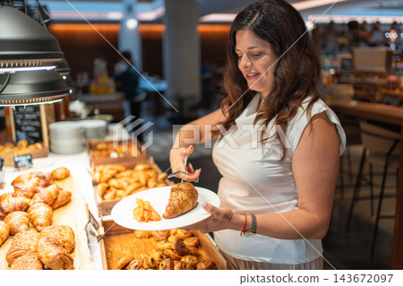 Woman chooses croissant, pancakes, salmon and cheese from a hotel breakfast buffet. Self service breakfast offers guests a variety of food during travel Woman chooses croissant, pancakes, salmon and cheese from a hotel breakfast buffet. Self service breakfast offers guests a variety of food during travel 143672097