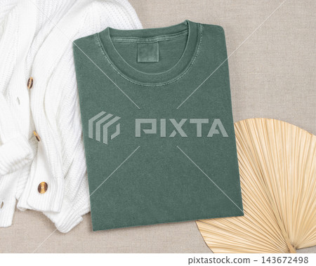 Washed green folded t-shirt mockup near knitted cardigan and palm leaf, template for print on demand 143672498