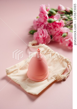 Reusable menstrual cup with cotton storage pouch and pink carnations on pink background Reusable menstrual cup with cotton storage pouch and pink carnations on pink background 143673085